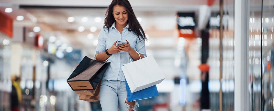 Brunette woman walking in a mall, holding shopping bags and staring at her smartphone.